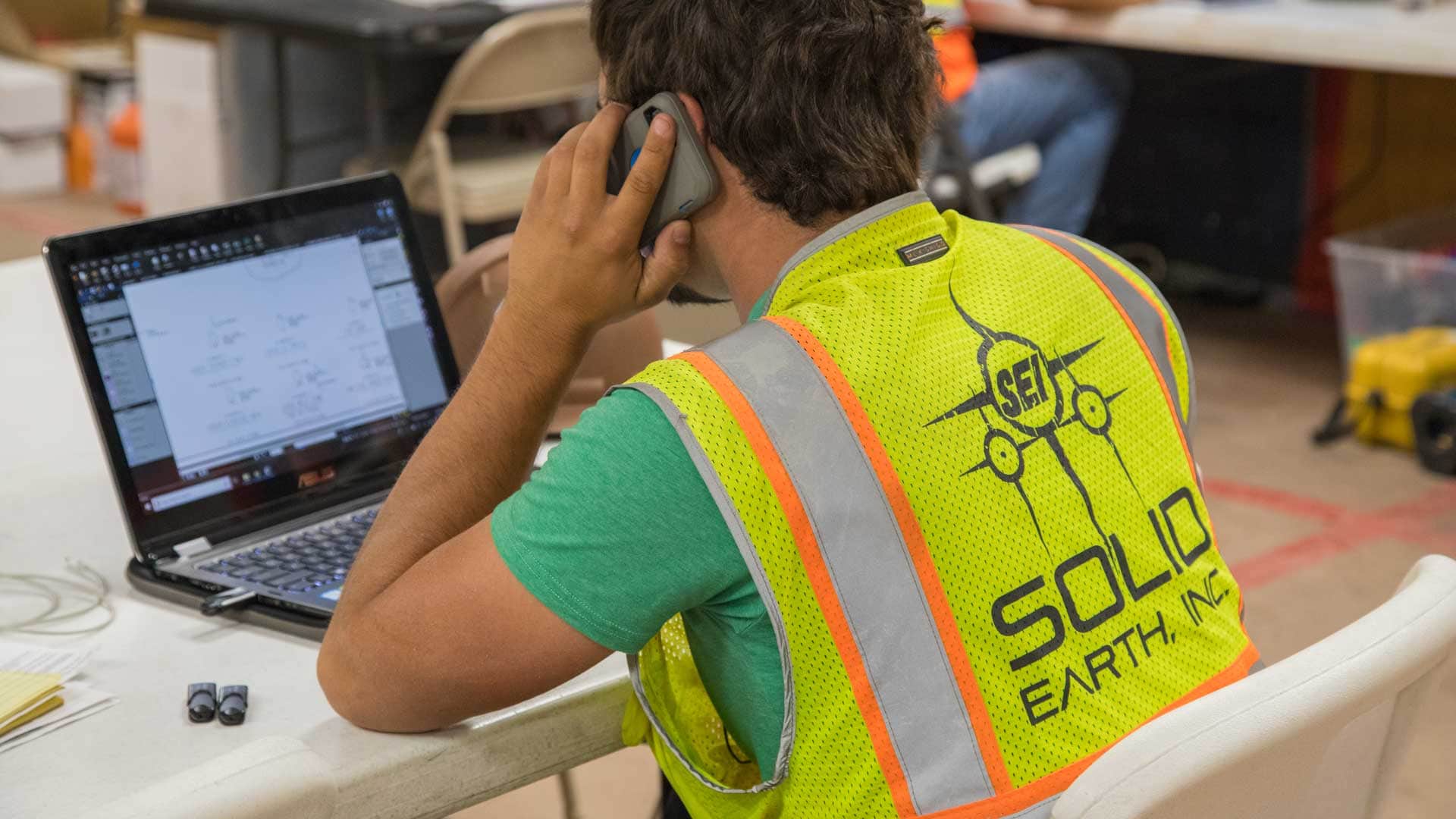 Man wearing Solid Earth Co safety vest working on a laptop indoors.